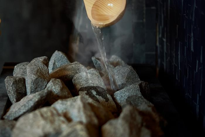 Wasser wird mit einer Holzschöpfkelle auf heiße Saunasteine gegossen, dabei steigt Dampf auf.