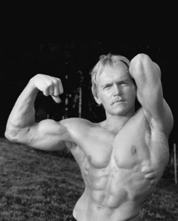 Ein muskulöser Mann posiert oberkörperfrei im Freien und zeigt eine Bodybuilding-Pose.