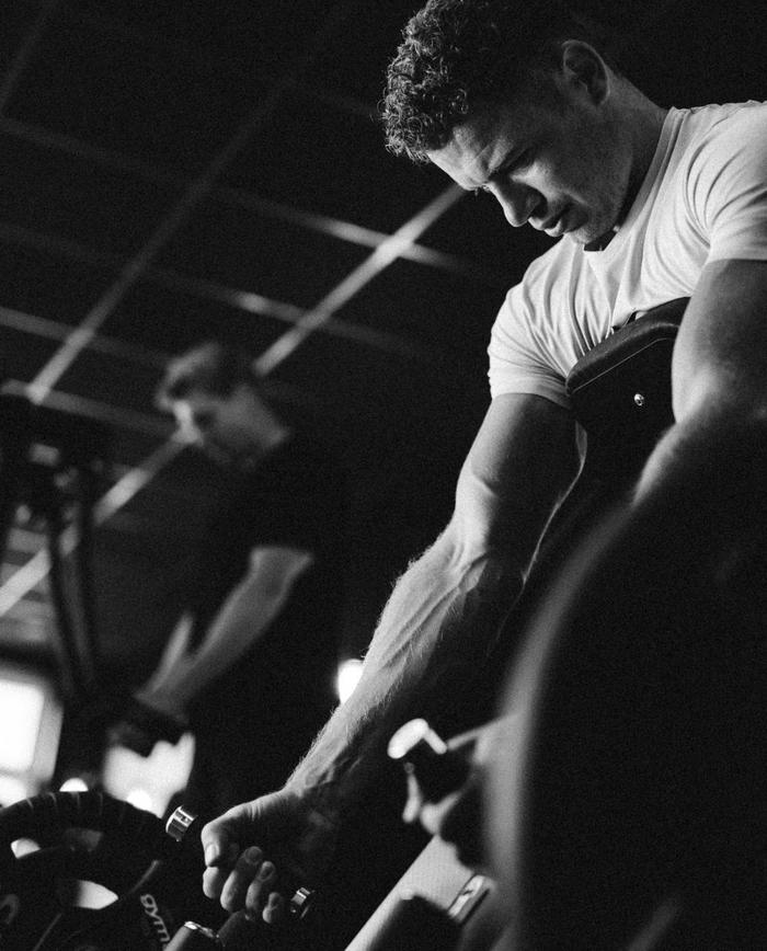 Ein Mann trainiert konzentriert an einem Fitnessgerät und zieht Gewichte nach oben; im Hintergrund trainiert eine weitere Person.