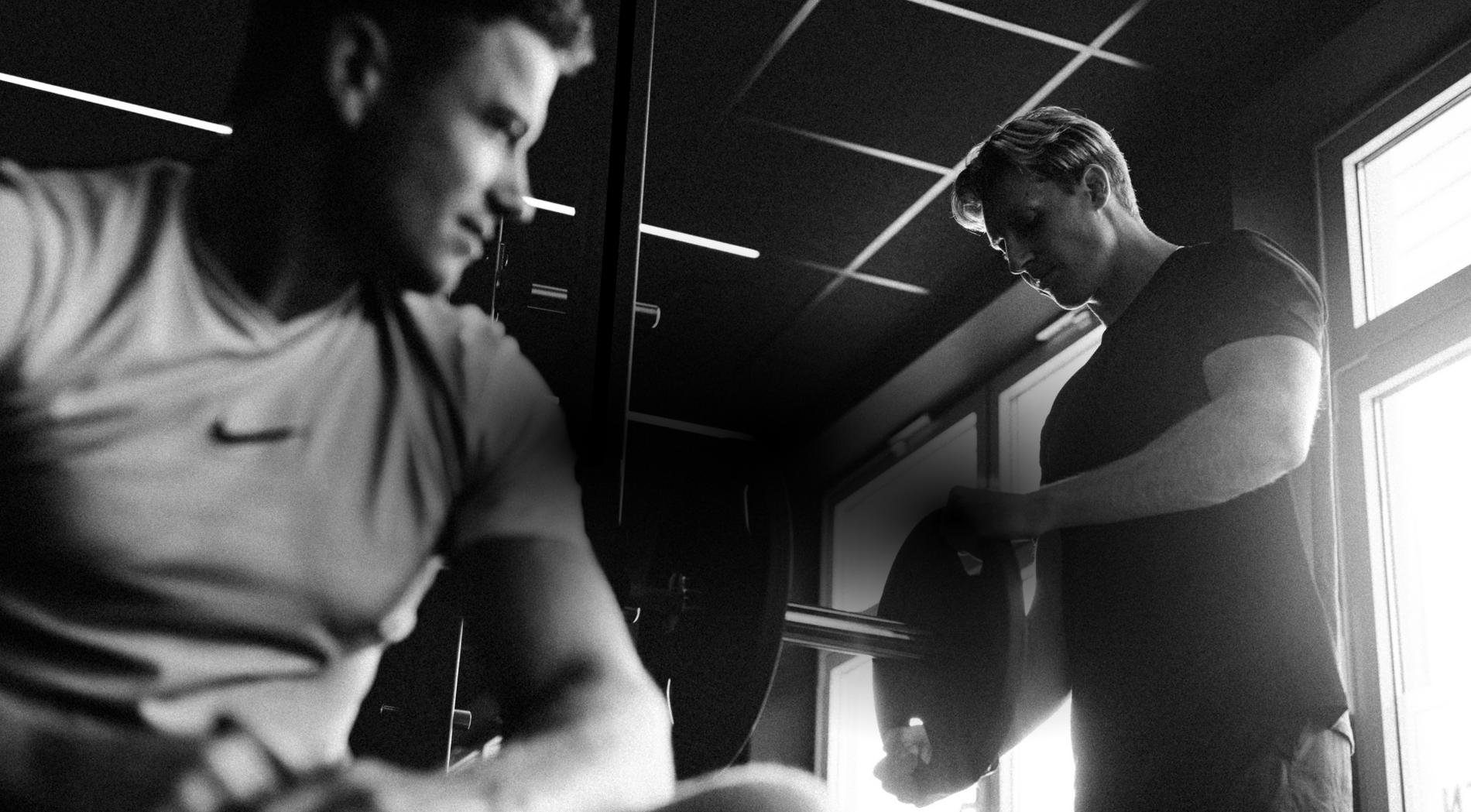 Zwei Männer im Fitnessstudio, einer sitzt im Vordergrund, der andere bereitet eine Hantel vor.
