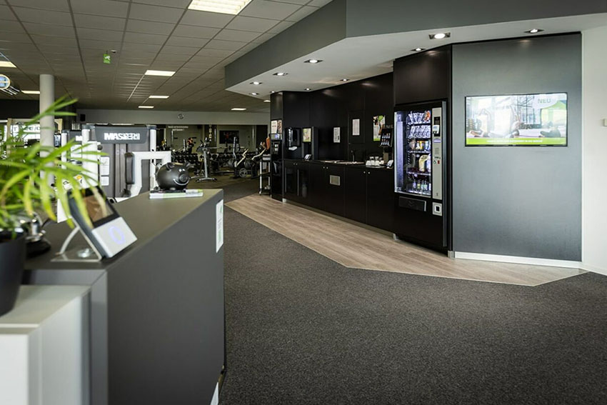 „Moderner Eingangs- und Aufenthaltsbereich eines Fitnessstudios mit Automaten und Blick in den Trainingsraum.