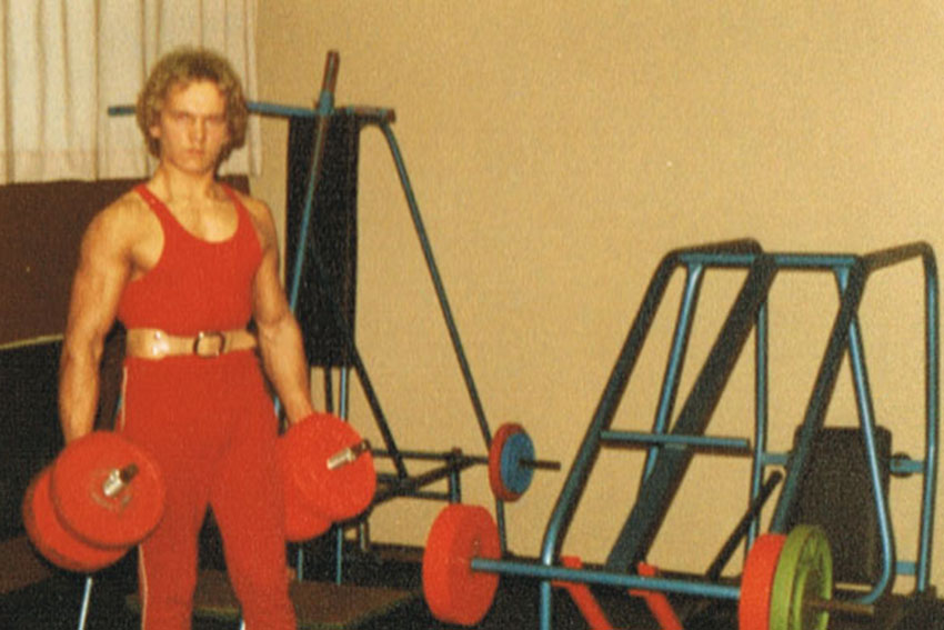 Historisches Foto eines jungen Mannes in roter Trainingskleidung mit Hanteln in einem alten Kraftraum mit einfachen Geräten.