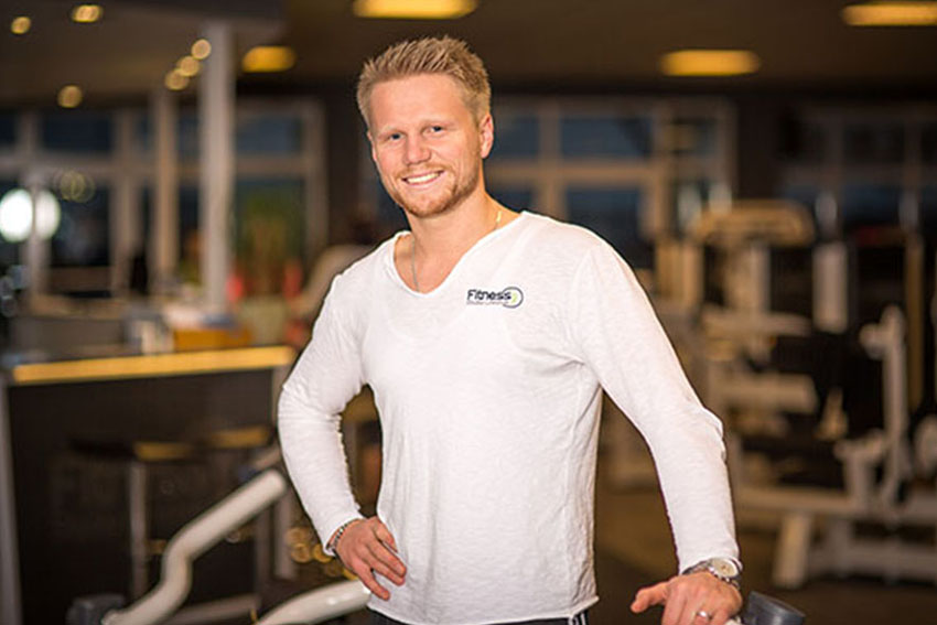 Lächelnder Mann in weißem Shirt lehnt in einem modernen Fitnessstudio an einem Trainingsgerät.
