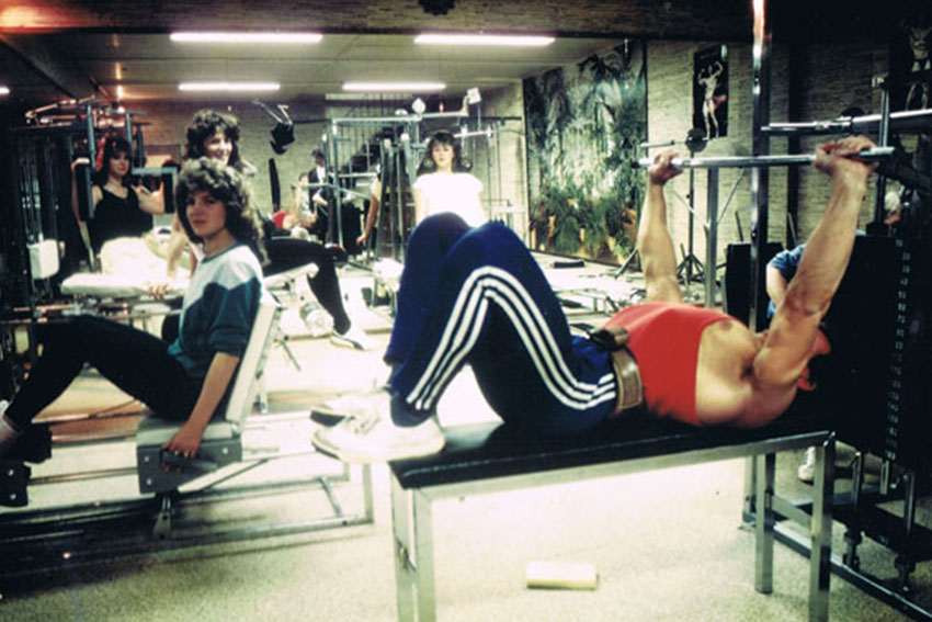 Gruppenszene in einem alten Fitnessstudio: Mehrere Personen trainieren an Geräten, im Vordergrund liegt ein Mann auf einer Bank und hebt eine Hantelstange.