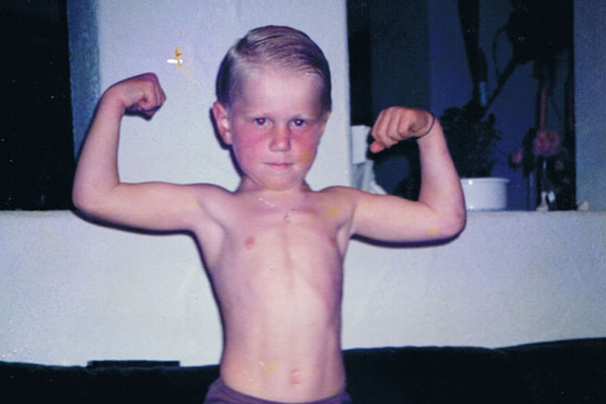 Junger Junge ohne Shirt steht im Wohnzimmer und zeigt lachend eine Muskelpose.