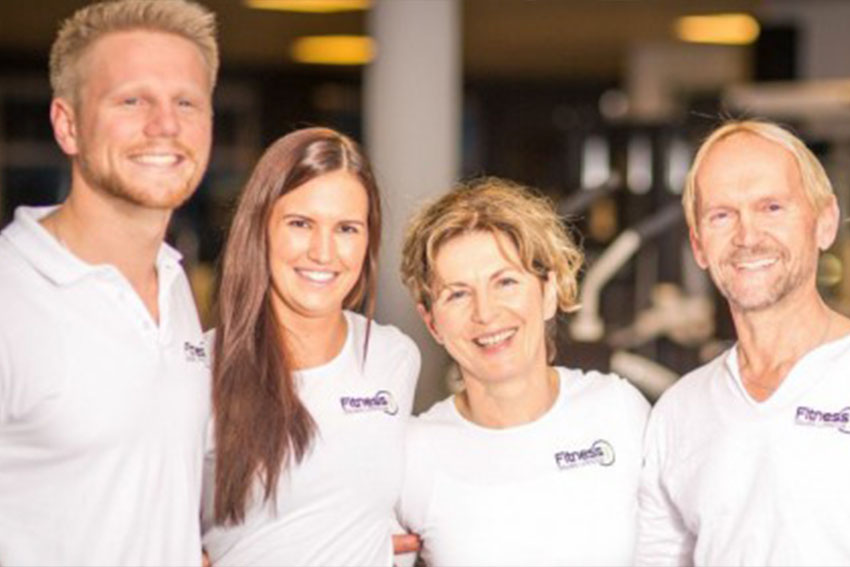 Vier lachende Teammitglieder in weißen Fitnessstudio-Shirts stehen zusammen für ein Gruppenfoto.