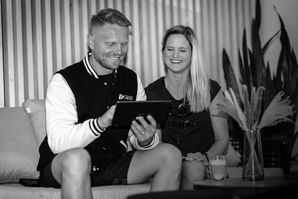 Ein Mann und eine Frau sitzen auf einem Sofa, lächeln und schauen gemeinsam auf ein Tablet, das der Mann in den Händen hält.