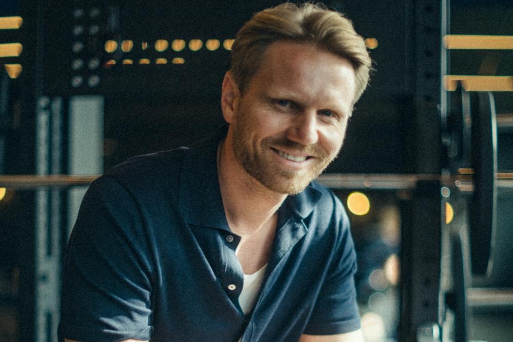 Mann mit blondem Haar lächelt in einem Fitnessstudio in die Kamera.